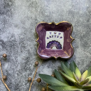 May include: A purple ceramic trinket dish with gold trim and the words "YOU MATTER" inside. The dish features a rainbow design and is a decorative item.
