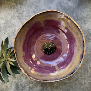 May include: A handmade ceramic bowl with a unique design. The bowl features a deep purple interior with a gold rim and a black center dotted with gold accents. The bowl's irregular shape adds to its artistic appeal. A decorative piece for the home.