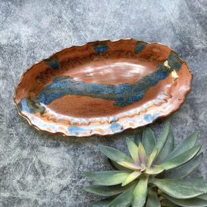 May include: Handmade ceramic serving platter. The oval-shaped dish features a brown base with blue and white accents. The rim has a wavy, organic edge. The platter is approximately 12 inches long and 6 inches wide.