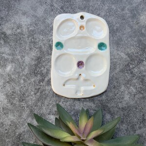 May include: White ceramic paint palette with five circular wells and a small bird design. The wells have colorful accents in green, purple, and orange.