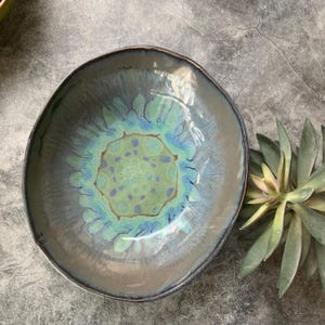 May include: A handmade ceramic bowl with a unique, organic shape. The bowl features a blend of colors, including shades of green, blue, and brown. The interior has a central design with a speckled pattern. The rim has a dark, metallic finish.