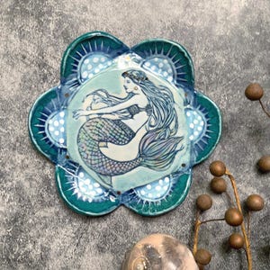 May include: A ceramic mermaid-themed decorative plate. The plate is flower-shaped with a teal rim and a central image of a mermaid with long flowing hair and a scaled tail. The mermaid is depicted in shades of blue, green, and purple.