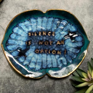 May include: A decorative ceramic dish with a blue and teal glaze and gold trim. The dish is shaped like a leaf and features the words "SILENCE IS NOT AN OPTION!" in gold lettering. The dish is a unique home decor item.