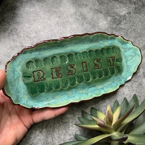 May include: A handmade ceramic tray with a rectangular shape and a wavy edge. The tray is glazed in shades of green and turquoise, with the word "RESIST" in red lettering. The tray is a unique piece of art.