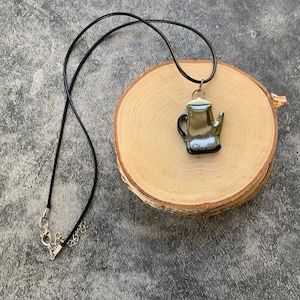 May include: A black cord necklace with a silver and black glass teapot pendant. The pendant is hanging on a wooden surface.
