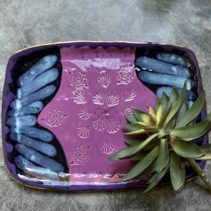 May include: Rectangular ceramic serving tray with a purple center and blue and black accents. The tray features a gold rim and a floral design in the center. A succulent plant is in the foreground.