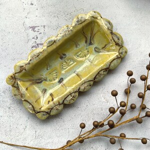 May include: A small, rectangular ceramic dish with a scalloped edge. The dish is glazed in a yellow and green color with a brown and white marbled pattern. The dish has a raised design of leaves on the bottom.