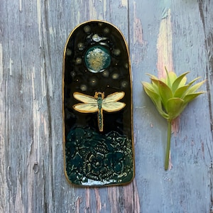 May include: A rectangular ceramic tray with a dark background, featuring a dragonfly design. The tray has a gold trim and a textured, dark teal base. The dragonfly is cream-colored with gold accents. A faux succulent is next to the tray.