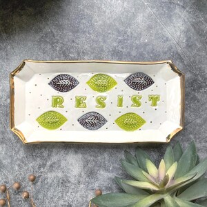 May include: Rectangular ceramic tray with gold trim and the word "RESIST" in green letters. The tray features green and dark blue leaf designs and small black dots on a white background. Decorative home decor.