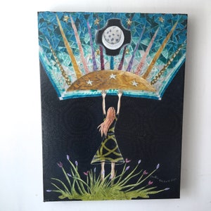 May include: A woman with long red hair is standing in a field of green grass and purple flowers. She is holding up a large, colorful mosaic piece that depicts a crescent moon and stars. The mosaic piece is set against a black background.