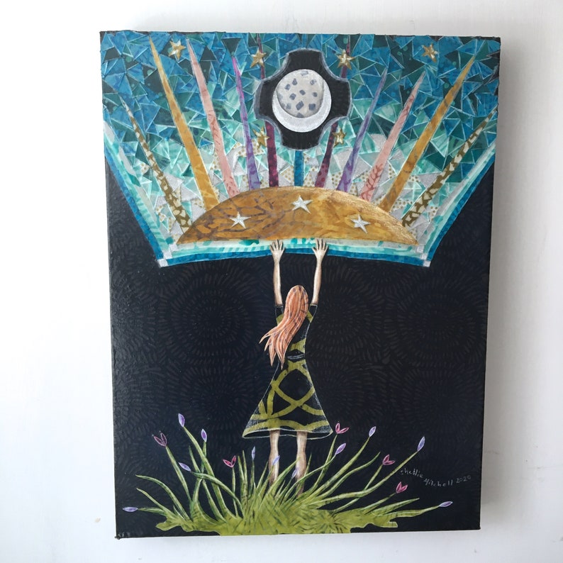 May include: A woman with long red hair is standing in a field of green grass and purple flowers. She is holding up a large, colorful mosaic piece that depicts a crescent moon and stars. The mosaic piece is set against a black background.