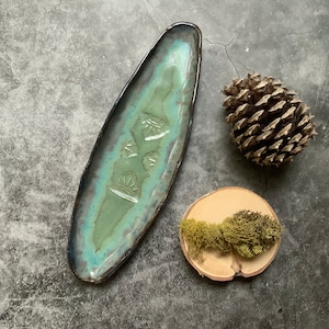 May include: A long, oval-shaped ceramic dish with a teal glaze and a black rim. The dish has a design of three stylized suns in a light brown color.