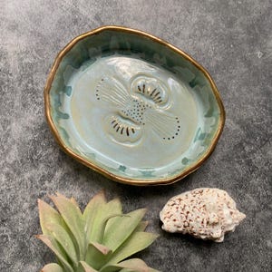 May include: A light blue ceramic dish with a gold rim and a raised floral design. The dish is oval shaped and has a glossy finish.