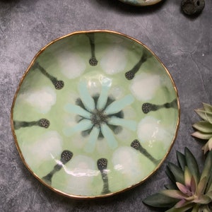 May include: A green ceramic bowl with a gold rim and a white and blue floral design. The bowl has a textured surface and is decorated with black dots.