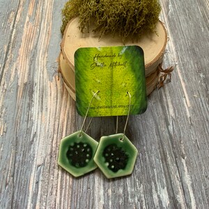 May include: A pair of green hexagonal ceramic earrings with silver hoops. The earrings have a dark green center with a white border. The earrings are handmade by Shellie Mitchell and are available on Etsy.