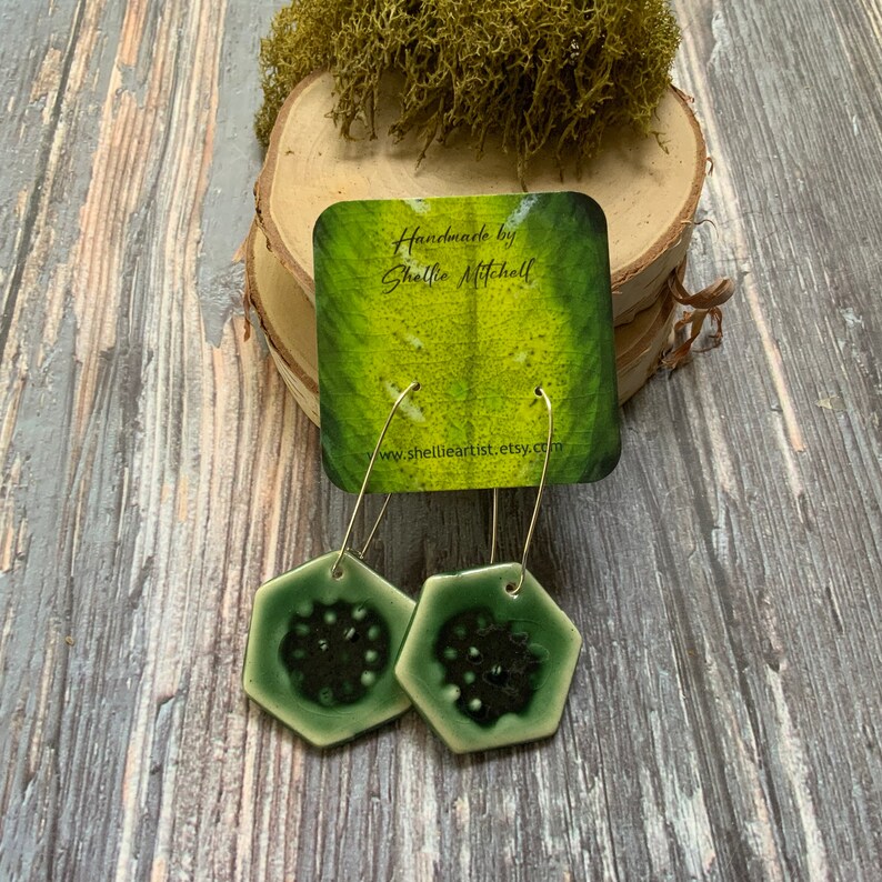 May include: A pair of green hexagonal ceramic earrings with silver hoops. The earrings have a dark green center with a white border. The earrings are handmade by Shellie Mitchell and are available on Etsy.