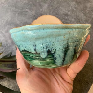 May include: A handmade ceramic bowl with a light blue glaze and green accents. The bowl has a unique, textured surface and a slightly rounded shape.
