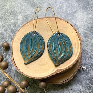 May include: A pair of teal and gold leaf-shaped earrings with gold-colored ear wires. The earrings are displayed on a wooden slice. The teal color has a textured appearance with gold-colored vertical lines.