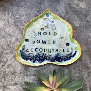 May include: A ceramic dish shaped like a leaf with a gold rim. The dish has a light blue background with a white bird flying above the words "HOLD POWER ACCOUNTABLE" in green and blue letters.