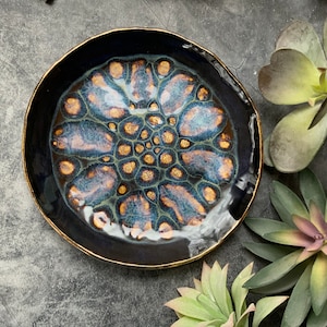 May include: A black ceramic plate with a gold rim and a blue and gold abstract design. The design resembles a flower with a circular pattern in the center.