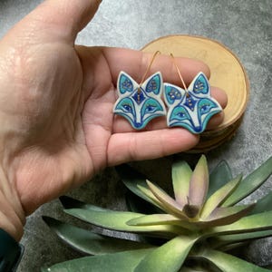 May include: A pair of ceramic fox earrings with blue, white, and gold details. The earrings are dangle style with gold hooks.