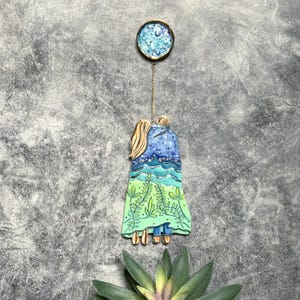 May include: A unique, handmade pendant featuring two figures embracing, with a blue and green landscape design. The pendant is suspended from a gold chain and topped with a round, blue and gold accent. The design evokes a sense of connection and nature.