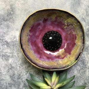 May include: A round ceramic bowl featuring a glossy glaze with a deep black center speckled with tiny white dots, surrounded by a vibrant magenta ring that transitions into a golden-yellow rim. The bowl has an organic, slightly irregular shape.