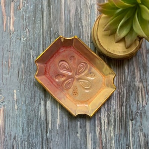 May include: A small, rectangular ceramic dish with a gold rim and a raised, stylized flower design in the center. The dish has a red and orange glaze with a subtle metallic sheen.