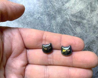 May include: A pair of black cat-shaped stud earrings with gold accents. The earrings are small and detailed, with a glossy finish. The cat faces have a simple, stylized design.
