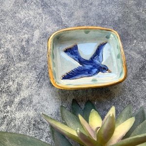 May include: A square ceramic dish with a blue bird design. The bird is painted in shades of blue with gold accents on the wings. The rim of the dish has a gold border. The background is a light blue color.