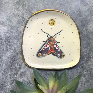 May include: A small, off-white ceramic dish with a gold rim. The dish features a hand-painted moth with red, black, and yellow details. Small black dots are scattered around the moth. A yellow sun-like design is above the moth.