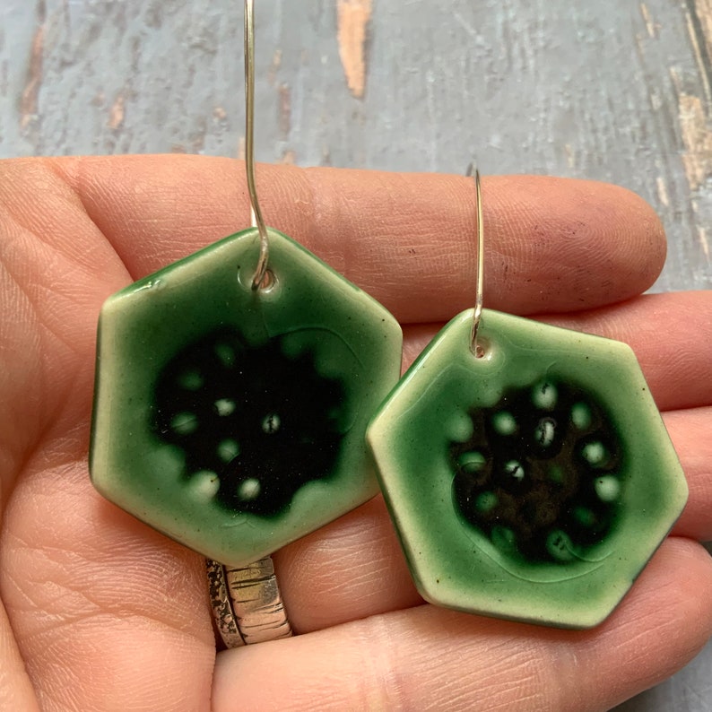 May include: A pair of green ceramic earrings with a hexagonal shape and a black and white design. The earrings are hanging from silver hooks.