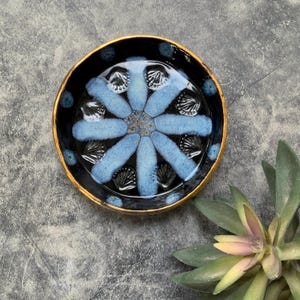 May include: A small, round ceramic dish with a blue and white floral design. The dish has a black background and a gold rim.