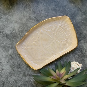 May include: A rectangular ceramic tray with a light pink glaze and a gold rim. The tray has a textured surface with a pattern of small circles.