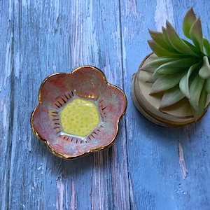 May include: A small, pink ceramic dish shaped like a flower with a yellow center. The dish has a gold rim and is decorated with a white and gold pattern.