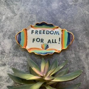 May include: A ceramic dish with a gold rim and the words "FREEDOM FOR ALL!" in black. The dish has a white base with colorful rainbow accents. A decorative succulent plant is in the foreground.