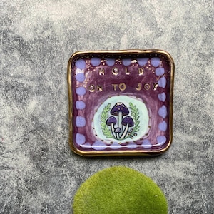 May include: A square, purple ceramic dish with a gold rim. The dish features a central design of three purple mushrooms on a light green background, surrounded by leaves. The words "HOLD ON TO JOY" are written in gold.