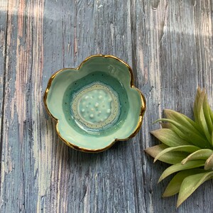 May include: A small, light blue ceramic dish with a gold rim and a white and blue patterned design. The dish is shaped like a flower.