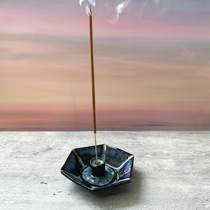 May include: A hexagonal ceramic incense holder with a glossy dark blue and black glaze featuring a central cylindrical stand. A single thin brown incense stick is inserted into the center, emitting a visible trail of white smoke upwards.