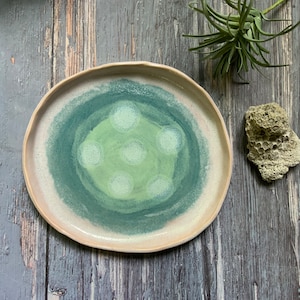 May include: A handmade ceramic plate with a unique design. The plate features a light pink rim, transitioning to a teal and green center with white dot accents. The plate's irregular edge adds to its handcrafted appeal.