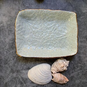 May include: A light blue ceramic serving dish with a textured surface and gold trim. The dish is rectangular and has a slightly raised edge. The dish is empty and is sitting on a gray surface.