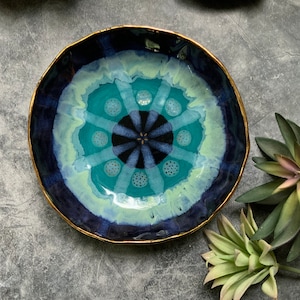 May include: A blue and green ceramic bowl with a gold rim and a mandala design. The bowl has a white background with a blue and green mandala design in the center. The mandala design has a black center with gold dots around it.