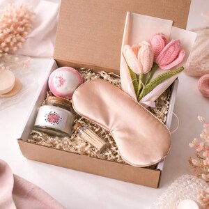 Op de afbeelding: Een geschenkdoos gevuld met zelfzorgartikelen. De doos bevat een roze slaapmasker, een kaars in een pot met de tekst "Love is in the air", een badbruisbal, een klein luciferflesje en een boeket gehaakte tulpen.