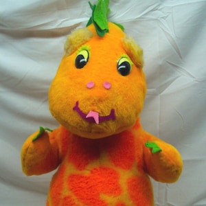 Può includere: Giocattolo di peluche arancione e giallo a forma di drago con foglie verdi sulla testa. Il giocattolo ha una lingua rosa che sporge e occhi neri.