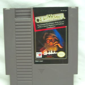 May include: A gray Nintendo Entertainment System (NES) game cartridge for the game "The Chessmaster." The game cartridge features a black and white image of a chessboard with chess pieces on it. The text "The Chessmaster" is printed in white on a black background. The text "The Ultimate Chess Partner - With 8 Levels of Play to Instruct Beginners and Challenge Grandmasters!" is printed in white on a red background.