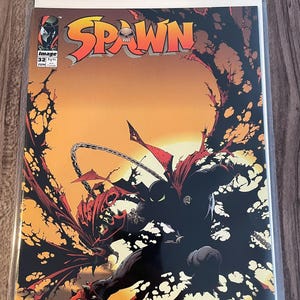 Pode incluir: Capa da banda desenhada Spawn com uma figura sombria com capa vermelha e manopla com espinhos, num fundo laranja e preto. O título "Spawn" é exibido em laranja e vermelho. O número 32 é visível.