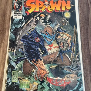 Op de afbeelding: Stripboekcover met de titel "SPAWN" in oranje en geel, met een schedel in de letter "A". De illustratie toont een grotesk clownachtig personage met een bloedig mes. Het stripboek is gelabeld met "Image" en "34".
