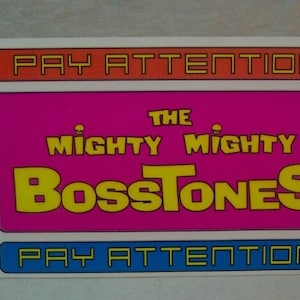 May include: A colorful poster with the text "Pay Attention" in orange, "The Mighty Mighty Bosstones" in yellow and pink, and "Pay Attention" in blue. The poster has a grid of images with people in various poses.