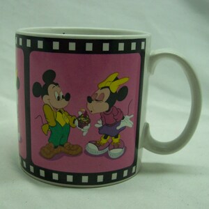 May include: White ceramic mug with a pink background and a black film strip border. The mug features a cartoon image of Mickey Mouse and Minnie Mouse. Mickey is holding a cupcake with a lit candle and Minnie is blowing out the candle.