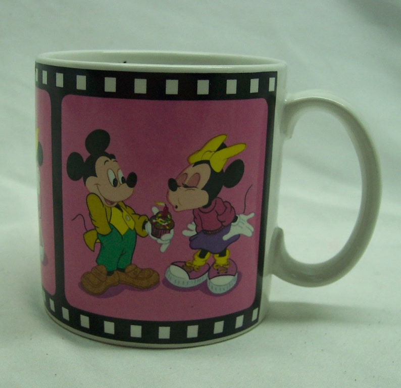 May include: White ceramic mug with a pink background and a black film strip border. The mug features a cartoon image of Mickey Mouse and Minnie Mouse. Mickey is holding a cupcake with a lit candle and Minnie is blowing out the candle.